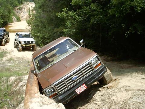 1985 Ford Bronco - A brown 1980s Ford Bronco navigates a challenging off-road course, tilting precariously in a ditch.