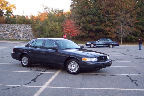 2001 Ford Crown Victoria - Two Ford Crown Victorias are parked in a lot during autumn, with a person standing near the rear vehicle.