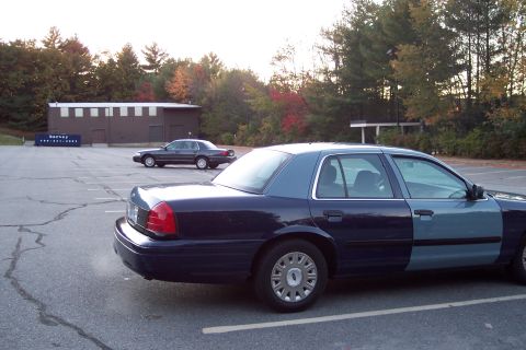 2001 Ford Crown Victoria - Two Ford Crown Victorias are parked in a lot with a building and trees in the background.