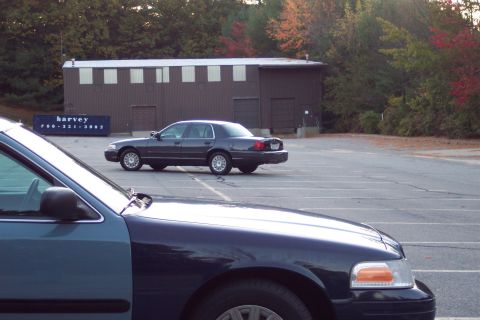 2001 Ford Crown Victoria - A dark sedan is parked in a lot, with another vehicle in the foreground. A dumpster with text is visible in the background.