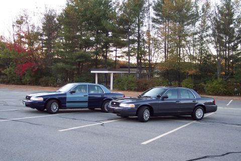 2001 Ford Crown Victoria - Two Ford Crown Victorias are parked in a lot, one with a two-tone paint job and the other in a solid dark color.