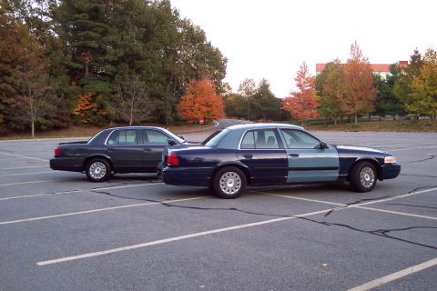 2001 Ford Crown Victoria - Two Ford Crown Victorias are parked in a lot with autumn trees in the background.