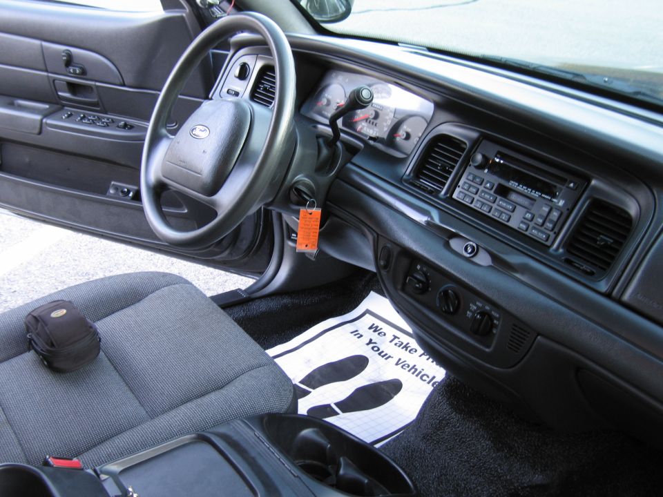 Interior of a 2001 Ford Crown Victoria showing the steering wheel, dashboard, radio, and driver's seat. Floor mat says 'We Take Pride In Your Vehicle'