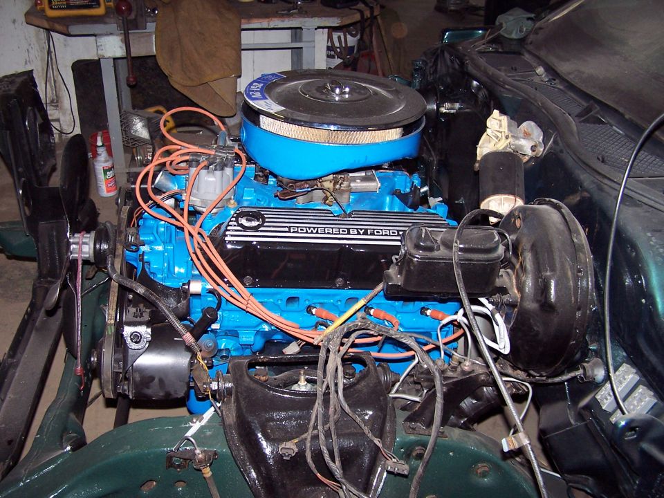 Blue Ford V8 engine with 'POWERED BY FORD' valve cover and orange plug wires in a garage.