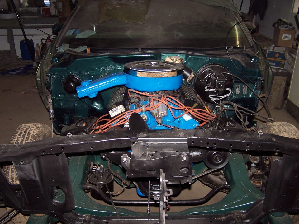 Green Ford engine bay with V8, blue air cleaner and orange plug wires, front end removed in garage
