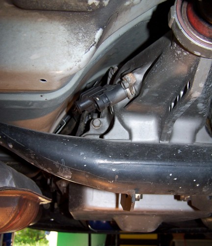 2000 Ford Crown Victoria - Underneath view of a vehicle's drivetrain, showing a sensor attached to the transmission housing. A black metal bar runs diagonally across…