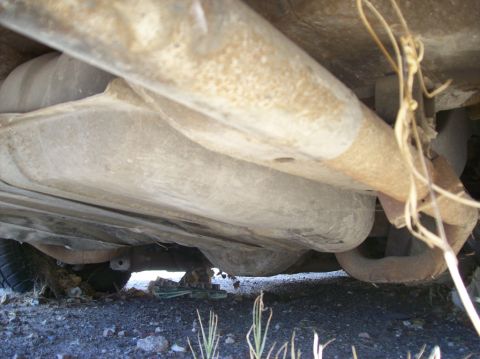 1975 Ford Ranchero - The underside of a vehicle shows the fuel tank and exhaust system. The vehicle appears to be resting on gravel with some dry vegetation vis…