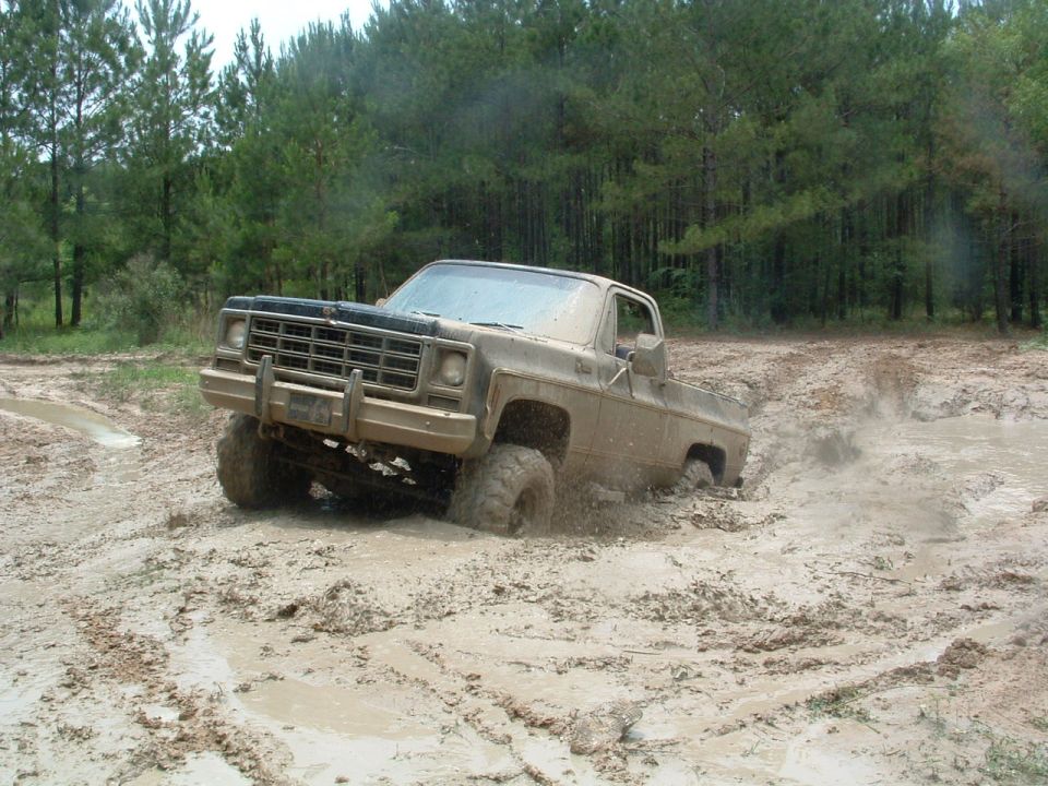 Tan Chevrolet pickup truck driving through deep mud with tires submerged and mud spraying.