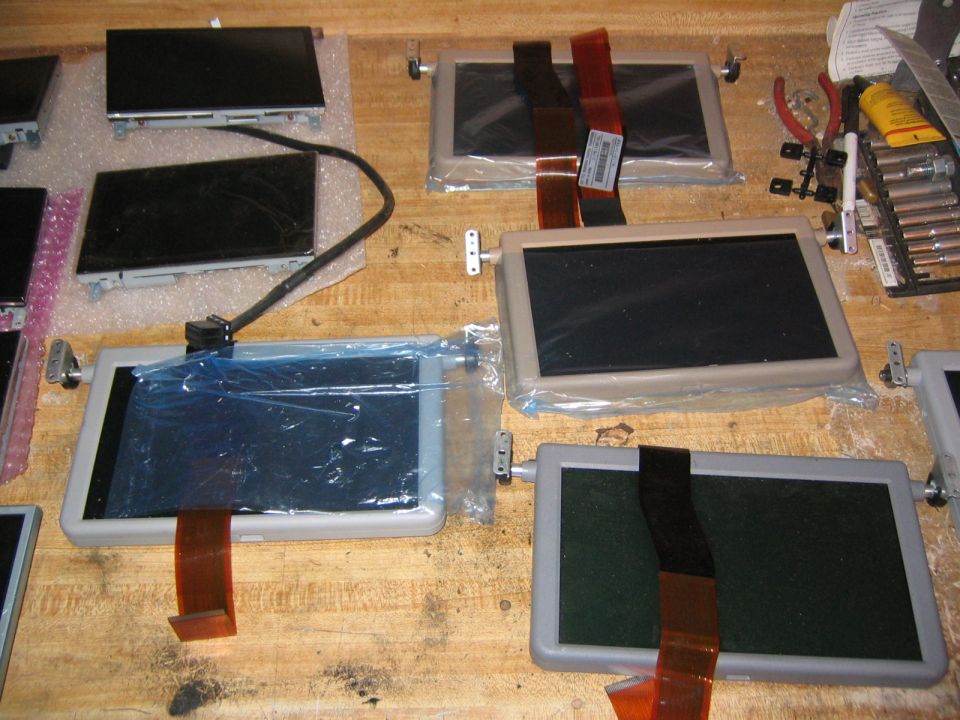 Multiple small electronic displays laid out on a wooden surface, some in plastic wrap with ribbon cables.