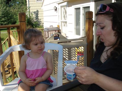 2000 Ford Crown Victoria - A woman is holding a container of yogurt and a spoon while sitting on a deck. A young child is sitting in a white chair in front of her.