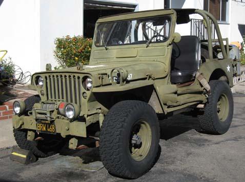 1948 Jeep Wrangler - A vintage olive green Jeep CJ-2A is parked on a paved surface in front of a building. The Jeep has large off-road tires, a winch mounted on…