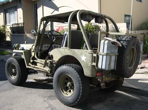 1948 Jeep Wrangler - The rear 3/4 view of a vintage olive drab Jeep CJ-2A is parked on a paved surface in front of a residential building. A spare tire, a Jerry…