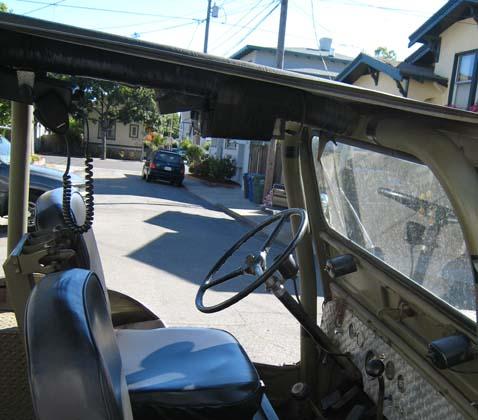 1948 Jeep Wrangler - Interior view of a vintage Jeep looking out the windshield at a street scene with a black SUV and houses. The Jeep has a black steering whe…