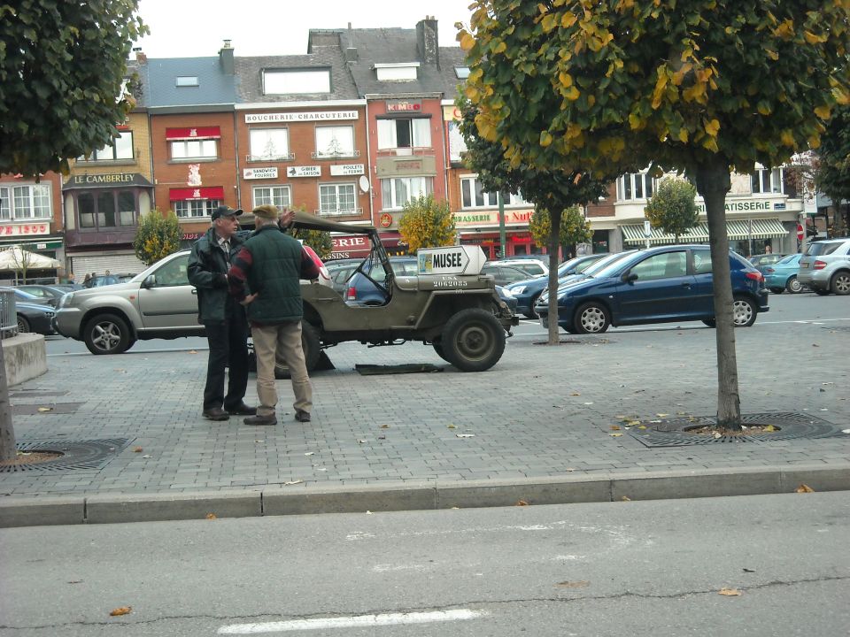 Olive green military jeep parked on cobblestones, with a sign reading MUSEE. Two men stand beside it. Other cars and buildings are in the background