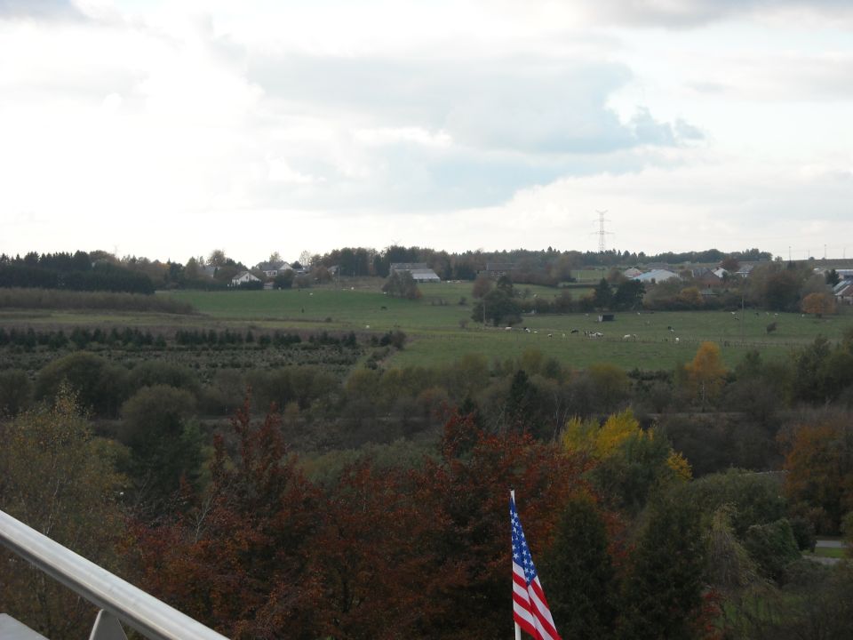 American flag in foreground with rural landscape of fields, trees, and buildings under a cloudy sky