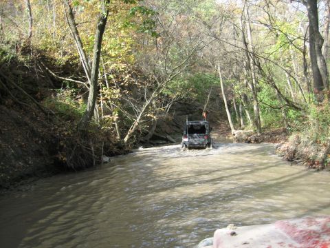 1991 Jeep Wrangler - A black Jeep Wrangler navigates a shallow river in a wooded area during autumn.