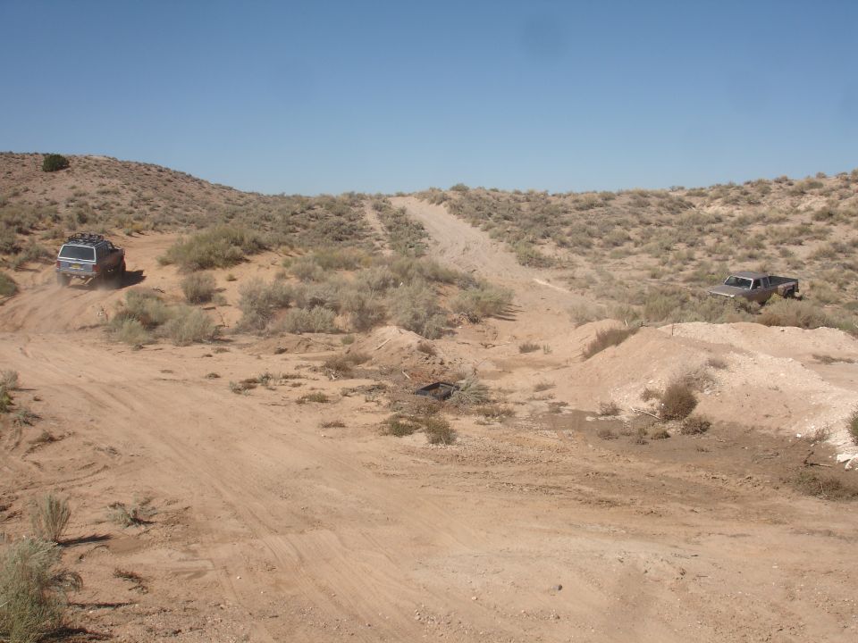 Two trucks driving on a dirt road in a desert landscape with scrub brush and hills