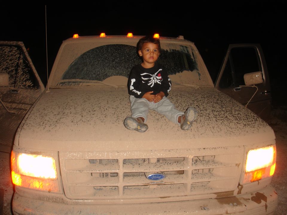 Kid in skeleton shirt sits on the hood of a dusty Ford truck. Another truck is visible to the left