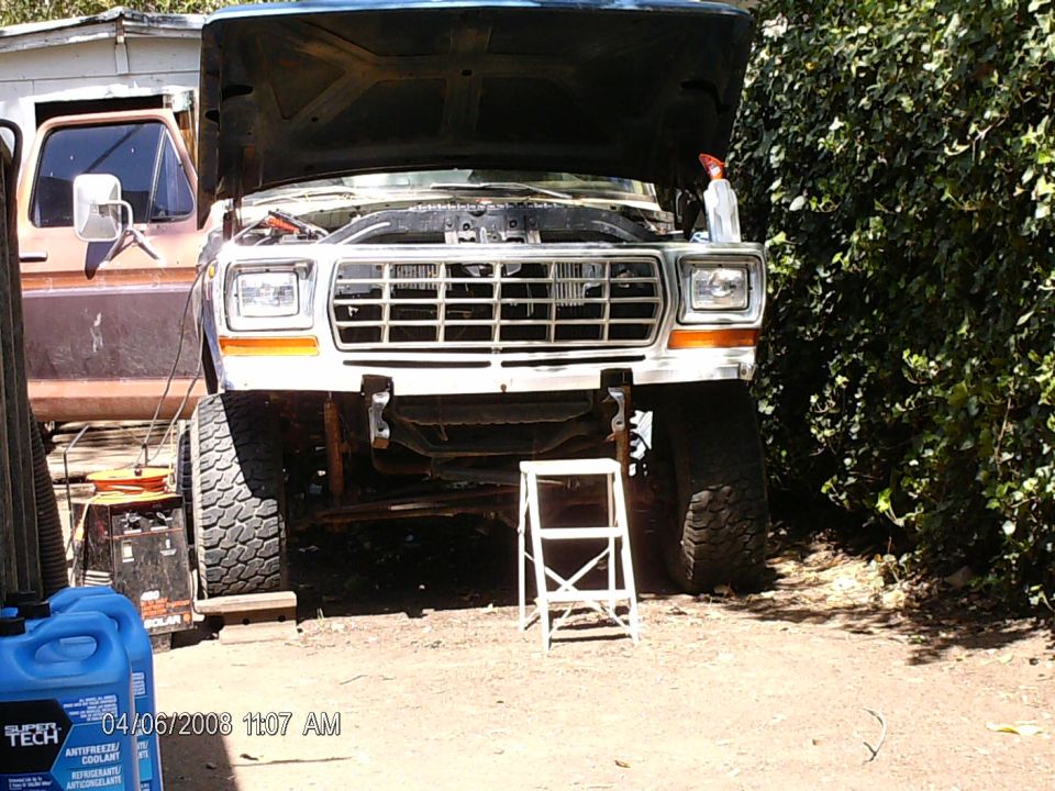 White 1979 Ford Bronco with hood open, chrome grille, parked on dirt next to a hedge and step stool