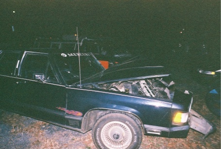 1988 Mercury Grand Marquis - The front end of a dark sedan is damaged, with the hood open to reveal the engine compartment.