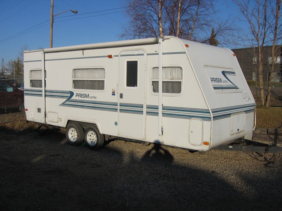 White Prism Ultra travel trailer with teal stripes, side view on gravel lot