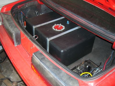 1988 Ford Bronco - Aftermarket fuel tank installed in the trunk of a red vehicle, with visible wiring and a SuperMotors.net watermark.
