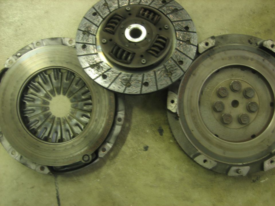 Three automotive clutch and flywheel components on a concrete surface. The parts are metallic and show signs of wear