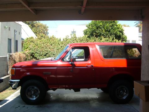 1988 Ford Bronco - Side view of a red SUV parked in a covered area with trees and buildings in the background.