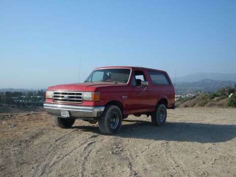 1988 Ford Bronco - A red Ford Bronco is parked on a dusty hill overlooking a town and distant mountains under a clear blue sky.