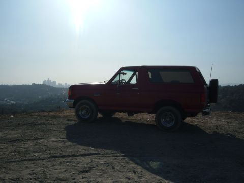 1988 Ford Bronco - Red full-size Ford Bronco in profile on a dirt overlook, backlit by the sun with a hazy downtown skyline and hillsides behind it. The remov…