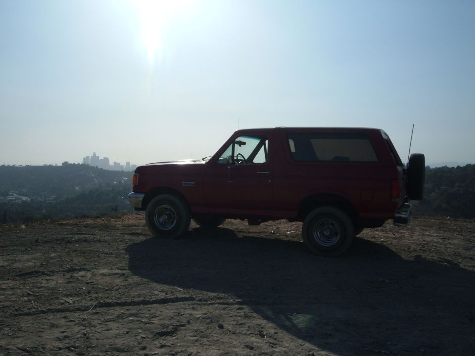 Red Ford Bronco in profile on a dirt overlook with city skyline in the haze behind