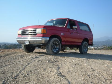 1988 Ford Bronco - A red first-generation-body Ford Bronco full-size SUV shown from a 3/4 front angle, parked on a dirt lot with hills in the background. It h…