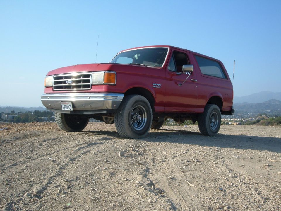 Red Ford Bronco SUV parked at 3/4 front angle on dirt lot with hills behind.