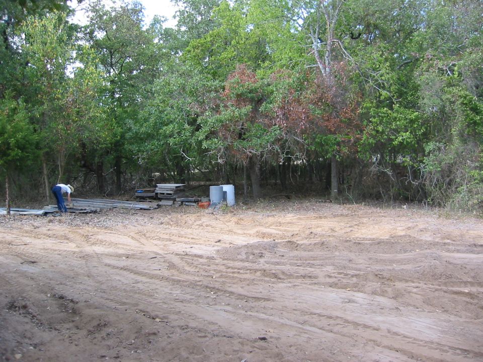 Person working near stacked lumber and concrete pipes in a dirt lot with trees in background