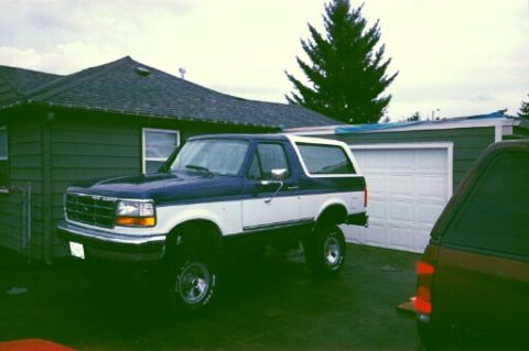 1996 Ford Bronco - A two-tone blue and white Ford Bronco is parked in front of a house and garage, with another vehicle partially visible on the right.