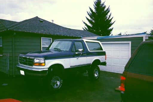 Two-tone blue and white Ford Bronco parked in a driveway next to a garage. The vehicle is lifted with aftermarket wheels