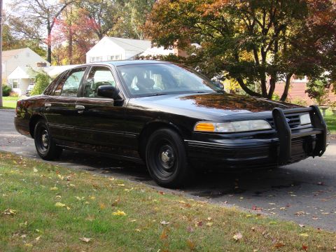 2003 Ford Crown Victoria - A black 2003 Ford Crown Victoria, equipped with a push bar, is parked on the side of a residential street.