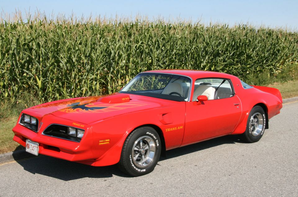 Red Pontiac Trans Am coupe with black hood bird decal parked beside a cornfield, front three-quarter view