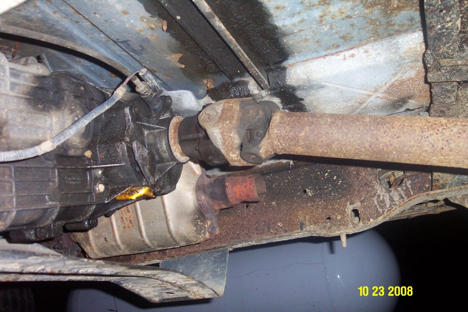 Undercarriage of a vehicle showing transmission, driveshaft, and exhaust system with rust and oil leaks