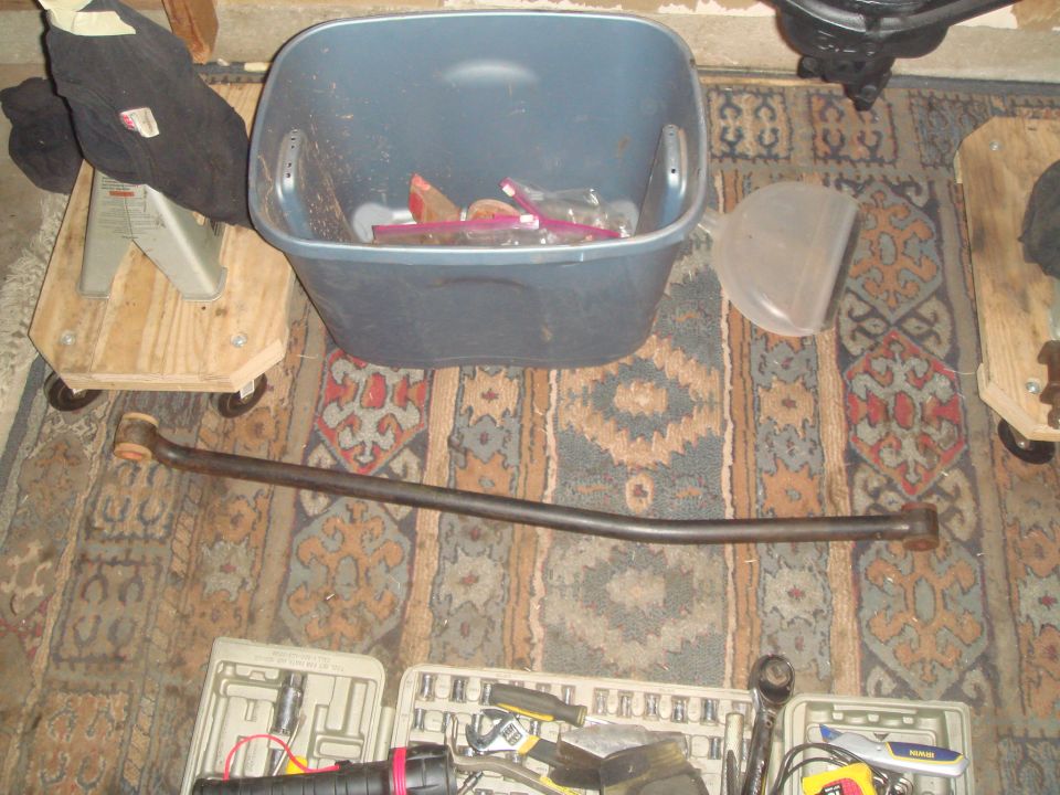 Curved metal automotive part with rubber ends, resting on a patterned rug near a blue bin and tools