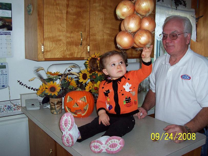 Child in Halloween sweater sits on kitchen counter next to a jack-o'-lantern and sunflowers, with a man standing nearby