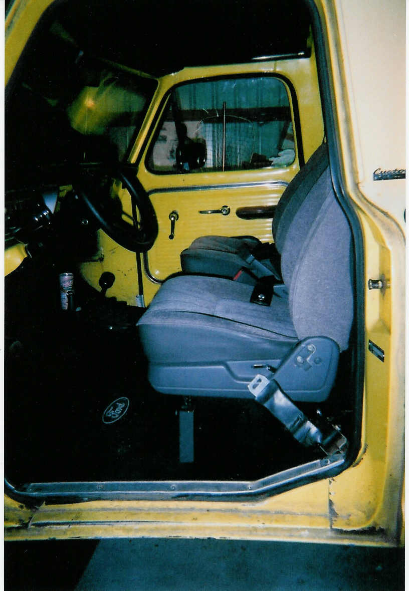 Open driver's door of yellow Ford truck showing gray bucket seat, steering wheel, and Ford floor mat