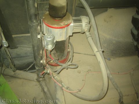 1990 Ford Bronco - cylindrical air filter and motor assembly with braided hoses and red wiring is mounted in the dusty engine bay of an off-road race vehicle.