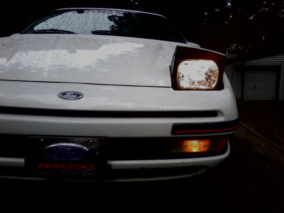 Front of white Ford at night with headlight on and 'RACING' plate.