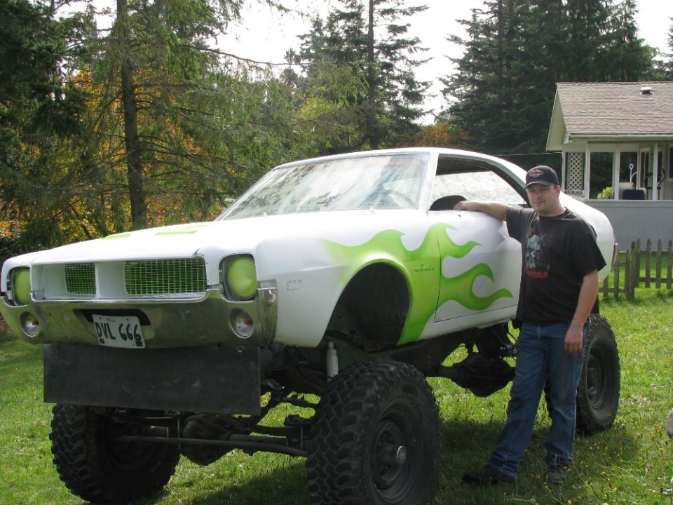 White coupe with green flames lifted on big off-road tires, man standing beside it