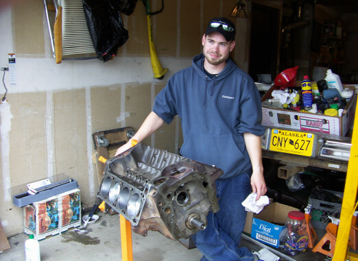 Man holding a bare engine block on a yellow stand in a garage