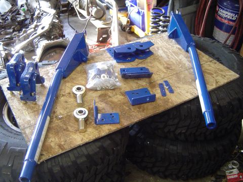 1979 Ford Bronco - Various blue-painted suspension parts and a bag of hardware are arranged on a piece of plywood resting on tires.