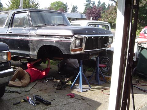 1979 Ford Bronco - Mechanics work on a black Ford Bronco lifted on jack stands in a driveway, with another truck visible in the background.
