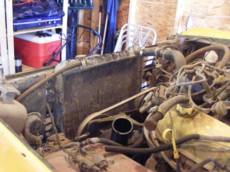 Engine bay of a yellow vehicle showing a dirty radiator, hoses, and wiring. A "SUPERMOTORS" watermark is visible