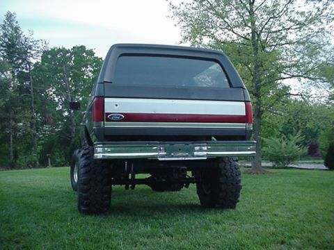1987 Ford Bronco - This lifted 1987 Ford Bronco is shown from the rear, parked on a grassy lawn with trees in the background.